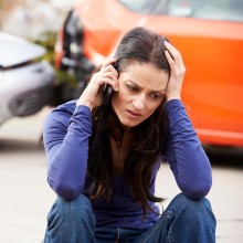 Frau sitzt am Boden und telefoniert, hinter ihr sieht man ein Unfallauto